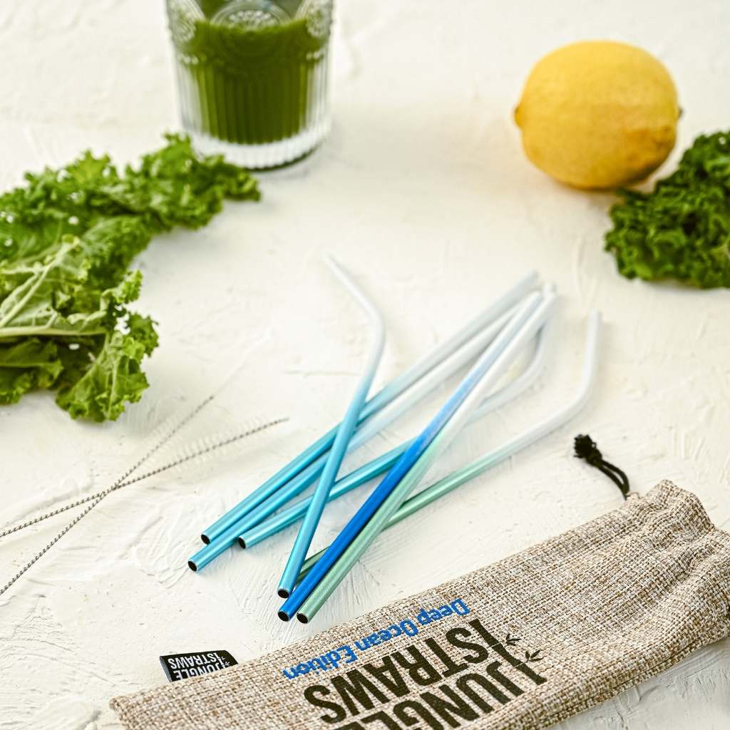 A collection of Jungle Culture - Stainless Steel Straws - Reusable Metal Straw Set: Living Coral Red alongside a lemon, a glass of green juice, and leafy greens, all resting on a pale surface with a burlap pouch reading "think green".