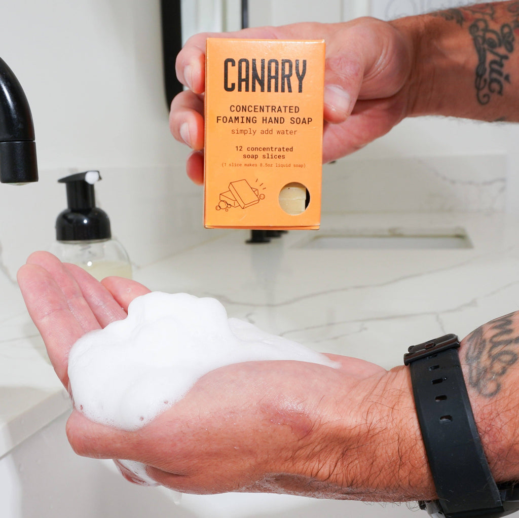 A person dispenses foam soap onto their hand from a sustainable, plastic-free concentrate packet labeled "Canary Clean Products - Cedar Mint Concentrated Hand Soap Refill Bar".