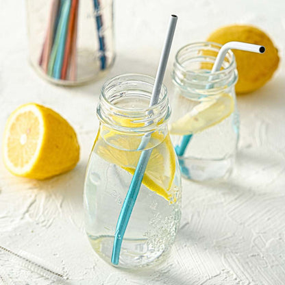 Two glass jars filled with lemon water, each with Jungle Culture stainless steel straws in deep ocean blue, accompanied by a half lemon, on a white textured surface.