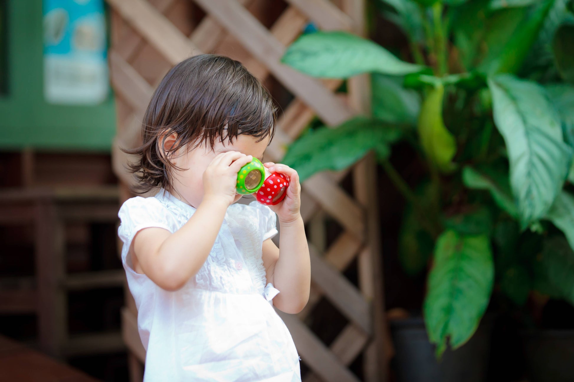 A young child exploring and blowing bubbles with a PlanToys Mushroom Kaleidoscope bubble wand outside near some vegetation.