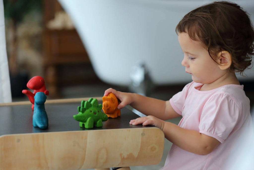 A young child is playing with a colorful, sustainable PlanToys Diplodocus figure on a wooden surface, focusing attentively on the toy.