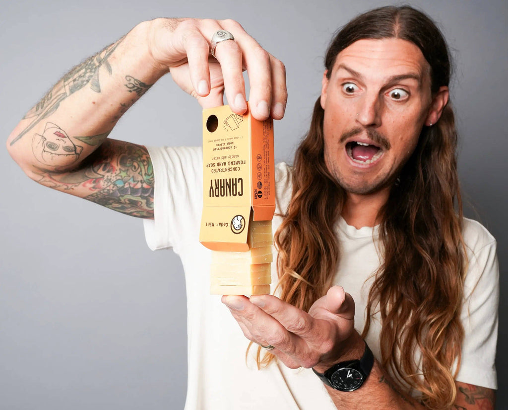 A surprised person with long hair and tattoos holding a giant, novelty cassette-shaped object made from sustainable Canary Clean Products - Cedar Mint Concentrated Hand Soap Refill Bars.