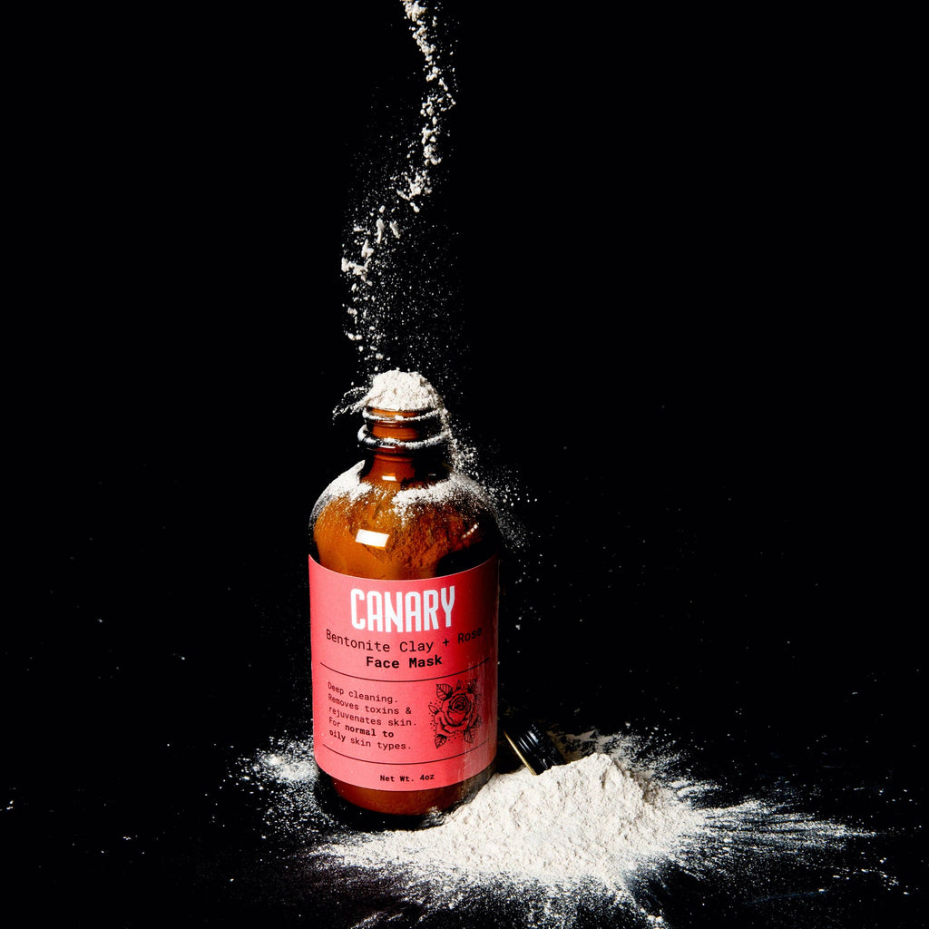 A bottle labeled "Canary Clean Products - Bentonite Clay + Rose Face Mask" with white powder being poured or spilling onto the surface around it against a dark background.