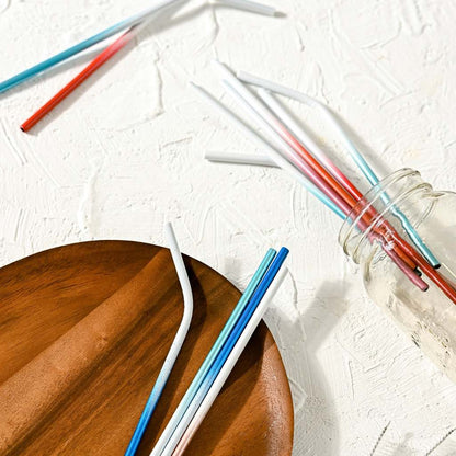 Assorted Jungle Culture stainless steel straws in Deep Ocean Blue are scattered on a textured white surface with some inside a glass jar, alongside a wooden plate with a straw resting on it. Shadows indicate bright, indirect light.