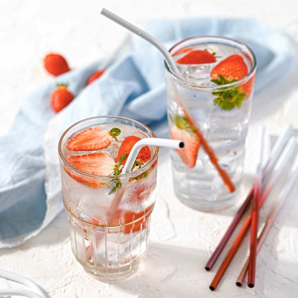 Two glasses of refreshing strawberry-infused water with mint leaves, accompanied by a blue napkin and colorful Jungle Culture - Stainless Steel Straws - Reusable Metal Straw Set: Deep Ocean Blue on a bright surface.