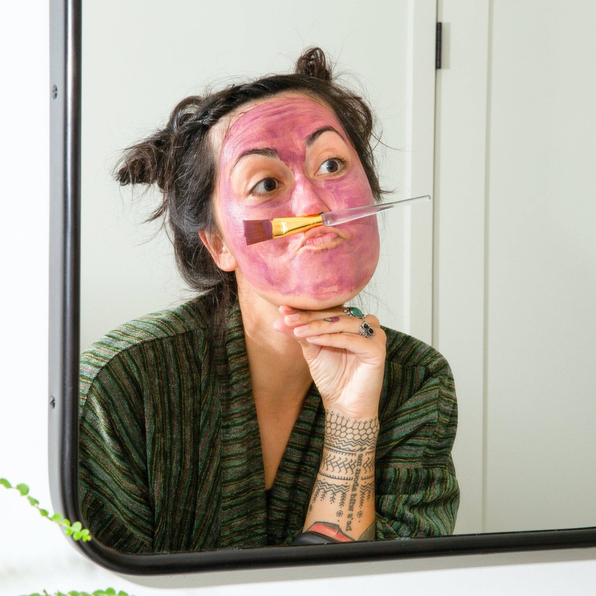 A person with a Canary Clean Products - Bentonite Clay + Rose Face Mask looking into a mirror, holding a cosmetic brush between their nose and upper lip, making a funny face.