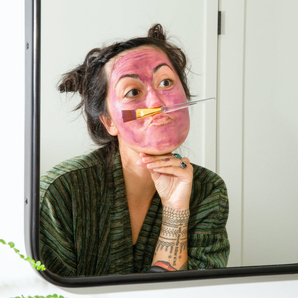 A person with a Canary Clean Products - Bentonite Clay + Rose Face Mask looking into a mirror, holding a cosmetic brush between their nose and upper lip, making a funny face.