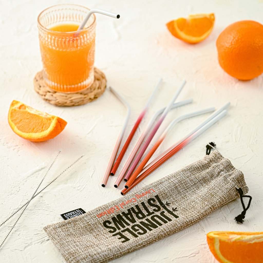 A glass of orange juice with a Jungle Culture - Stainless Steel Straws - Reusable Metal Straw Set: Living Coral Red sits beside fresh orange slices and a set of colored pencils on a textured surface, with a pencil case that reads "think less, live more.