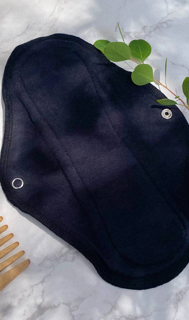 A navy blue Plum Thyme Moderate- Organic Reusable Menstrual Cloth Period Pad with snaps and leak protection, placed on a marble surface alongside a green leafy branch and a wooden comb.