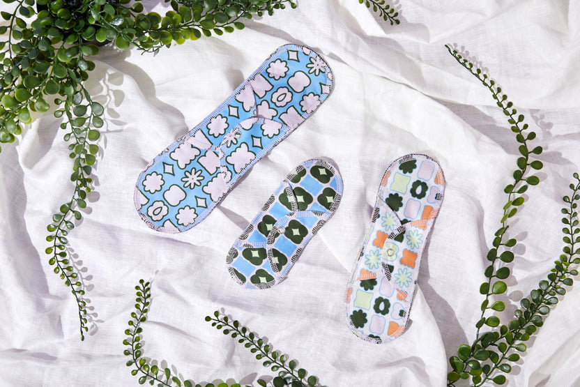 Two Plum Thyme Moderate- Organic Reusable Menstrual Cloth Period Pads, boasting enhanced absorbency, are placed on a crumpled white organic cotton fabric surface, surrounded by green leafy twigs.