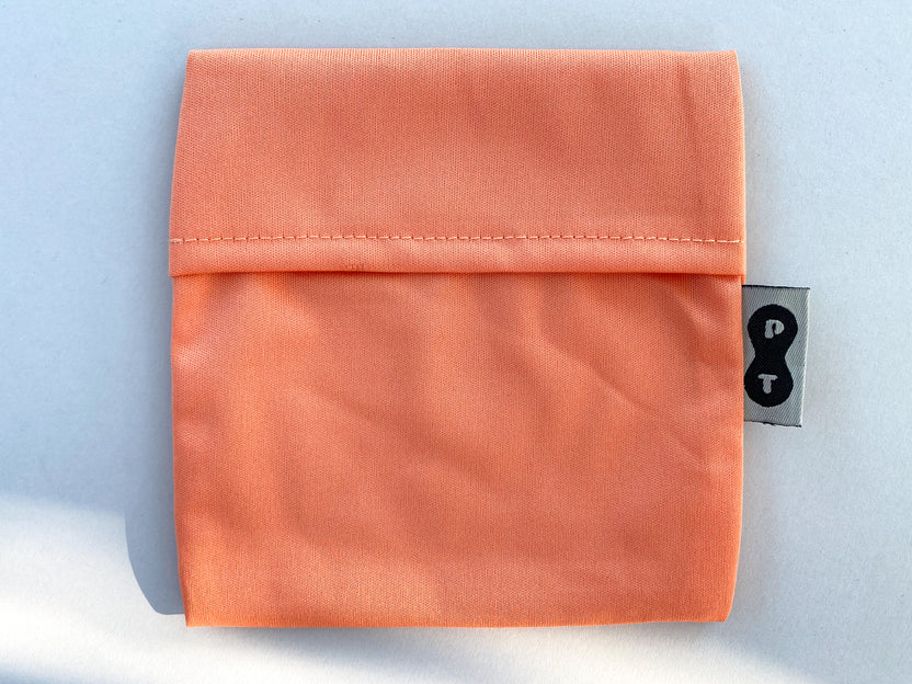An orange eco-PUL fabric pouch with a stitched seam and a small black tag featuring a white question mark, designed as a Plum Thyme pad wrapper.