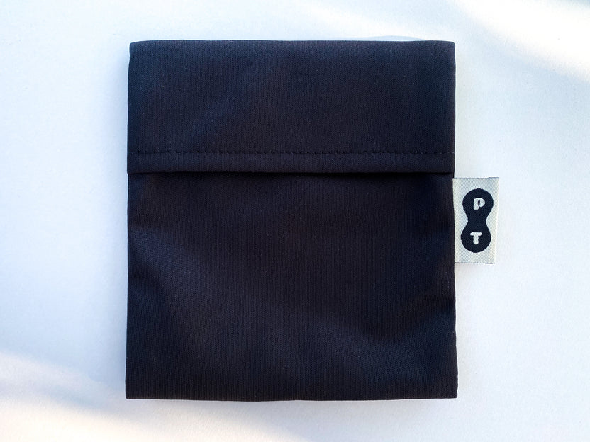 A folded black eco-PUL fabric Pad Wrapper by Plum Thyme with a tag displaying a white question mark and exclamation point on a gray background, designed as a mess-free pad wrapper.