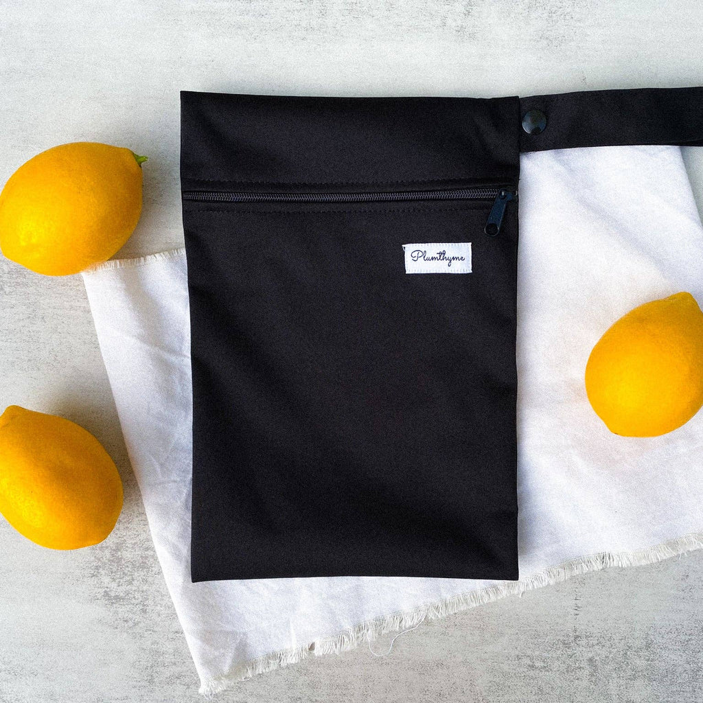 A black waterproof Plum Thyme PUL zippered pouch lying on a white cloth next to three bright yellow lemons.