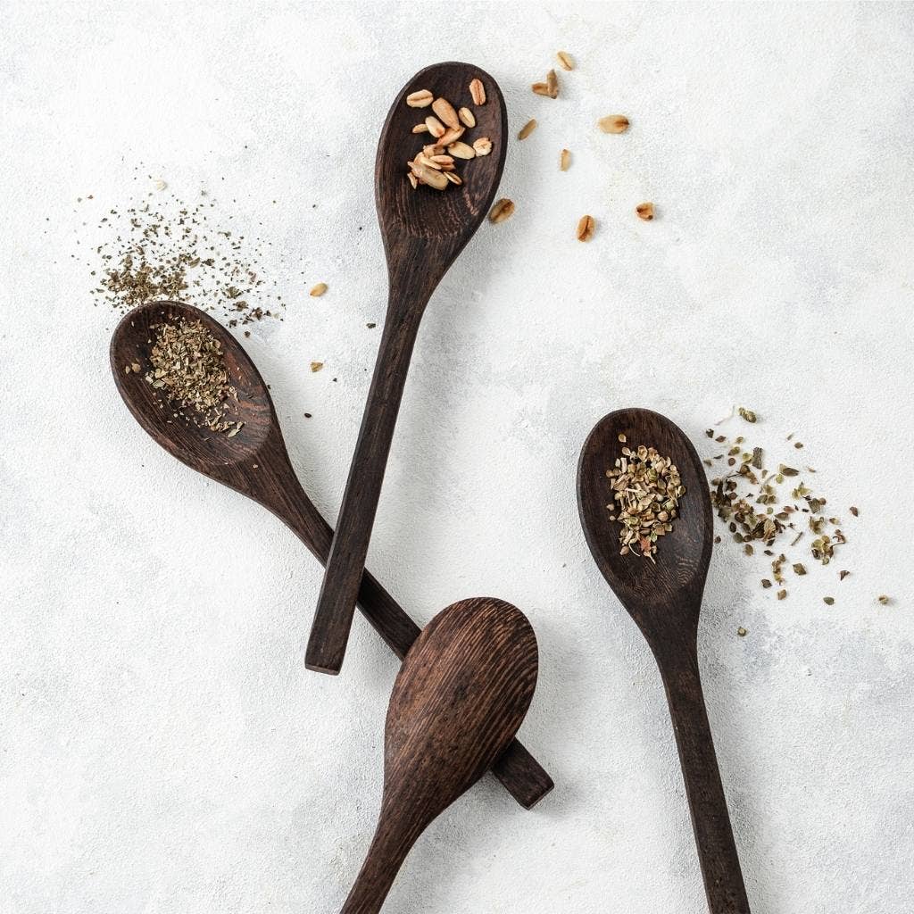 Four Jungle Culture Coconut Bowls Spoons on a textured surface, each containing different types and amounts of seeds and herbs, scattered lightly around them.