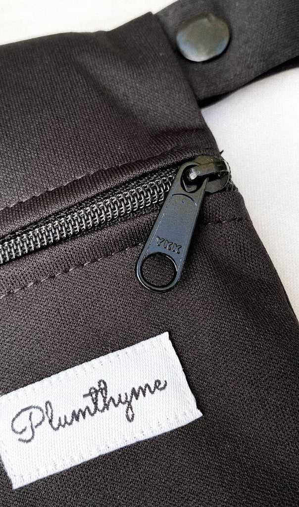 Close-up of a black waterproof Medium Wet Bag clothing item with a zipper partially unzipped, showing a brand label with the name "Plum Thyme".