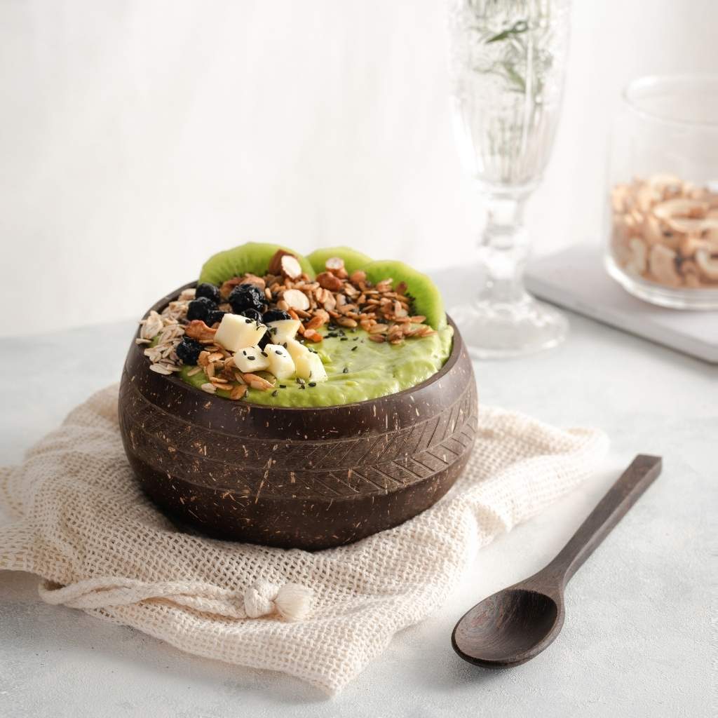 A smoothie bowl served in Jungle Culture's handmade Coconut Bowls garnished with kiwi slices, blackberries, granola, and nuts, accompanied by a Jungle Culture Coconut Bowls Spoon on a light-colored surface.