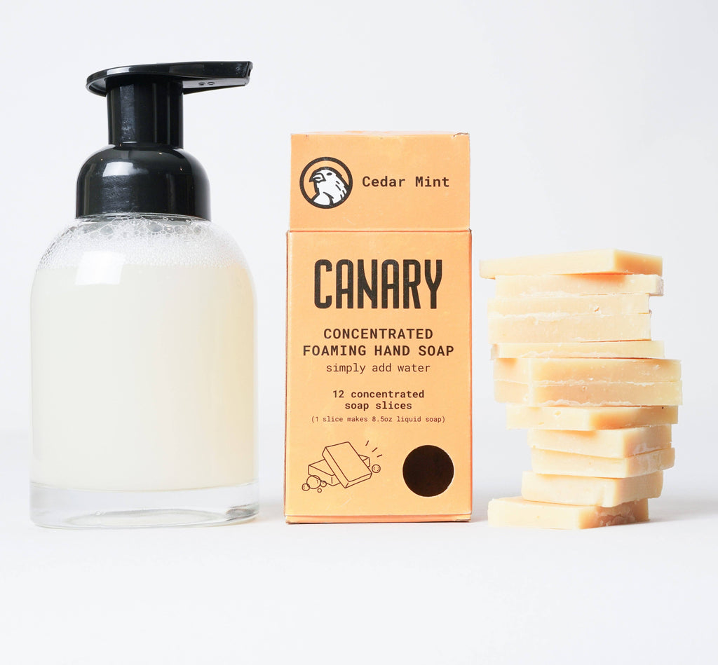A clear pump bottle with foaming hand soap next to a box labeled 'Canary Clean Products - Cedar Mint Concentrated Hand Soap Refill Bar' and a stack of yellow, plastic-free, sustainable hand soap refill bars.
