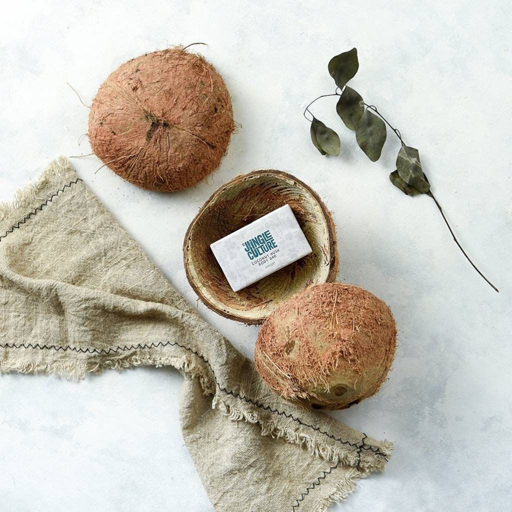 Two whole coconuts and one halved coconut displaying a bar of Jungle Culture's Natural Solid Bar Soaps (Coconut Husk) inside it, arranged on a textured cloth next to dried eucalyptus leaves on a light surface.